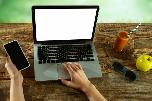 Écrans vierges d'ordinateur portable et de smartphone sur une table en bois à l'extérieur avec la nature sur le mur Fruits et jus de fruits frais à proximité. Concept de lieu de travail créatif, entreprise, indépendant. Copyspace.