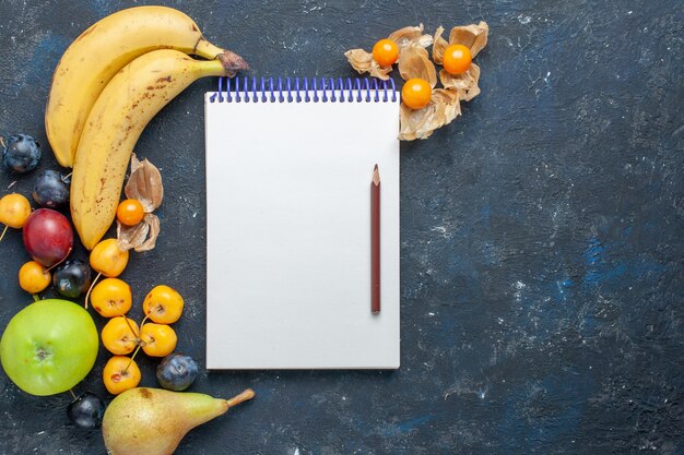 Crayon de bloc-notes de bananes jaunes vue de dessus avec pommes vertes fraîches, poires, prunes et cerises douces sur le bureau noir