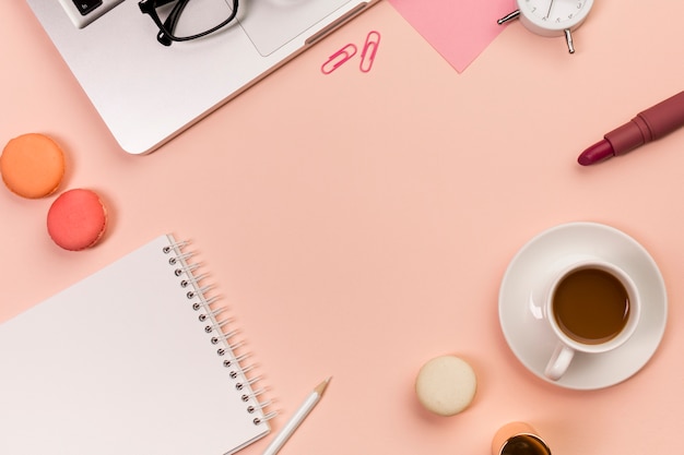 Crayon, macarons, bloc-notes à spirale, tasse à café, rouge à lèvres, lunettes de vue sur un ordinateur portable par-dessus le fond couleur pêche