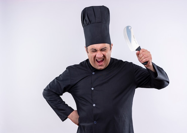 Crazy mad male chef cuisinier vêtu d'un uniforme noir et chapeau de cuisinier balançant un couteau criant avec visage en colère debout sur fond blanc