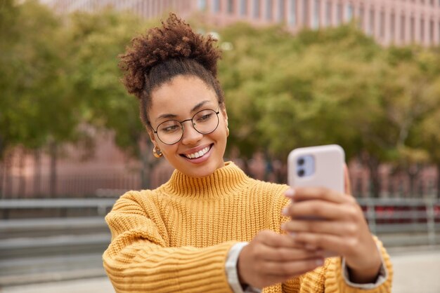 Un créateur de contenu positif du millénaire tire une influence vidéo vlog aime le mode de vie en réseau prend un selfie via un smartphone porte des lunettes rondes et un cavalier jaune pose sur un arrière-plan flou