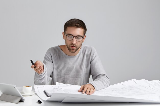 Un créateur masculin sérieux et concentré porte un pull ample et des lunettes rondes, regarde attentivement les croquis