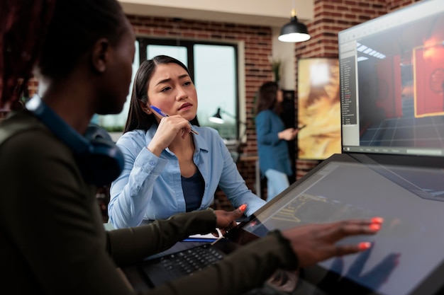 Créateurs de jeux créatifs multiethniques assis à leur bureau avec plusieurs écrans tout en travaillant avec des ressources 3D. Travailleurs de l'industrie du divertissement interactif numérique travaillant avec CGI.