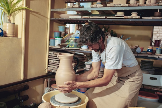 Création d'un pot ou d'un vase d'argile blanche en gros plan. Maître pot. Mains d'homme faisant macro de cruche d'argile. Le sculpteur dans l'atelier fabrique une cruche en gros plan en faïence. Tour de potier tordu.