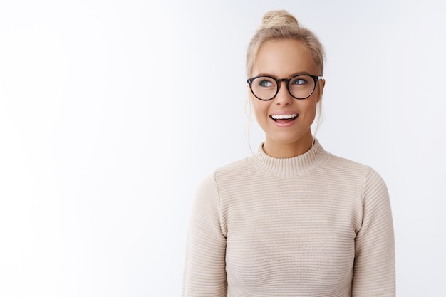 Créative et heureuse jolie blonde blonde caucasienne en pull et lunettes ayant une idée intéressante et géniale en regardant le coin supérieur gauche rêvasser souriant satisfait sur un mur blanc