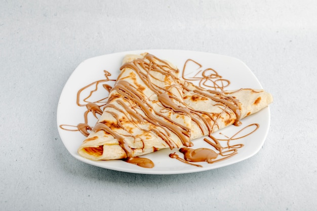 Crêpe au sirop de cacao au chocolat dans une assiette blanche sur un fond blanc.