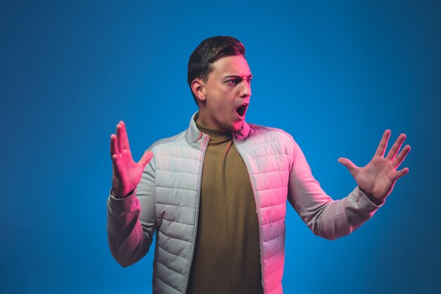 Des cris de colère. Portrait d'un homme de race blanche isolé sur un mur bleu en néon rose. Beau modèle masculin en casual. Concept d'émotions humaines, expression faciale Copyspace.