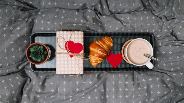 Photo gratuite croissant, coeur d'ornement, tasse de boisson, plante et coffret cadeau sur plateau