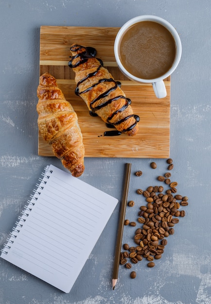 Croissants avec café, cahier, crayon, grains de café sur plâtre et planche de bois, vue de dessus.