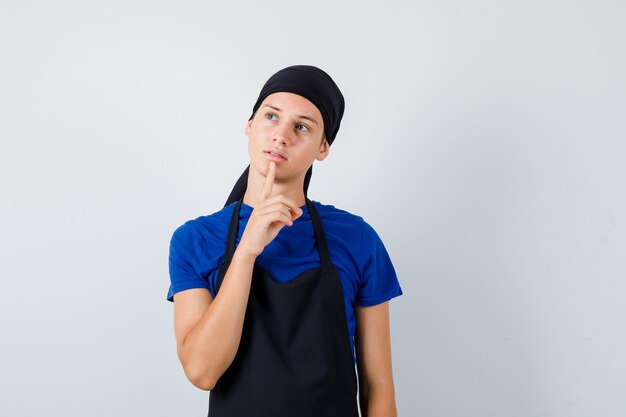 Cuisinier adolescent masculin gardant le doigt sur le menton en t-shirt, tablier et l'air préoccupé. vue de face.