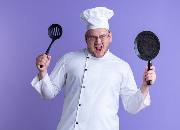 Cuisinier mâle adulte furieux portant un uniforme de chef et des lunettes montrant une cuillère à fentes et une poêle à frire à l'avant en regardant devant crier isolé sur un mur violet