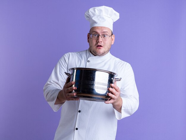 cuisinier mâle adulte surpris portant l'uniforme du chef et des lunettes regardant l'avant s'étirant vers l'avant du pot isolé sur un mur violet