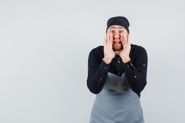 Cuisinier mâle disant secret avec les mains près de la bouche en chemise, tablier, vue de face.