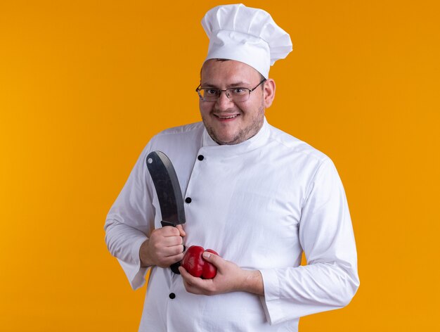 Cuisinier masculin adulte joyeux portant l'uniforme de chef et les verres tenant le couperet et le poivre regardant l'appareil-photo d'isolement sur le fond orange