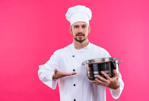 Cuisinier professionnel masculin en uniforme blanc et chapeau de cuisinier tenant une casserole vide présentant le bras de sa main souriant debout confiant sur fond rose