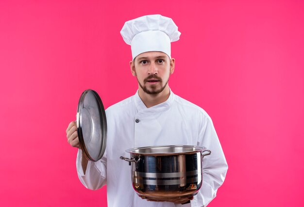 Cuisinier professionnel masculin en uniforme blanc et chapeau de cuisinier tenant une casserole vide regardant la caméra avec une expression confiante sérieuse debout sur fond rose