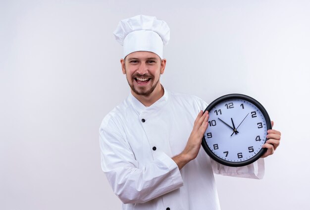 Cuisinier professionnel masculin en uniforme blanc et chapeau de cuisinier tenant une horloge regardant souriant joyeusement debout sur fond blanc