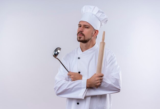 Cuisinier professionnel masculin en uniforme blanc et chapeau de cuisinier tenant la louche et le rouleau à pâtisserie regardant la caméra avec une expression suspecte debout sur fond blanc