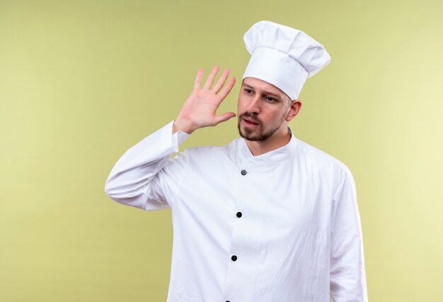 Cuisinier professionnel masculin en uniforme blanc et chapeau de cuisinier tenant la main près de son oreille en essayant d'écouter la conversation sur quelqu'un debout sur fond vert