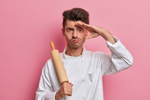 Un cuisinier sérieux tient un rouleau à pâtisserie, garde la main sur le front, réfléchit à ce qu'il faut cuisiner pour les visiteurs du restaurant