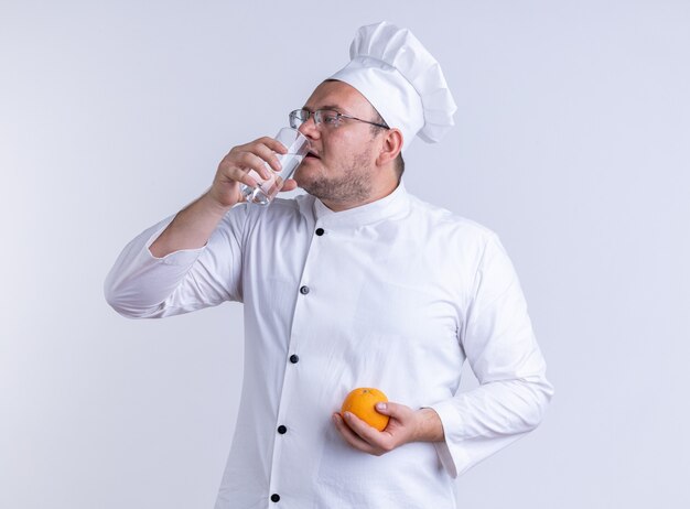 Cuisinier de sexe masculin adulte portant l'uniforme du chef et des lunettes tenant l'orange regardant le côté buvant un verre d'eau isolé sur un mur blanc