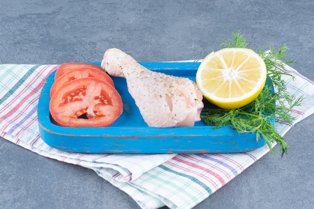 Cuisse de poulet non cuite, tranche de tomate et de citron sur plaque bleue.