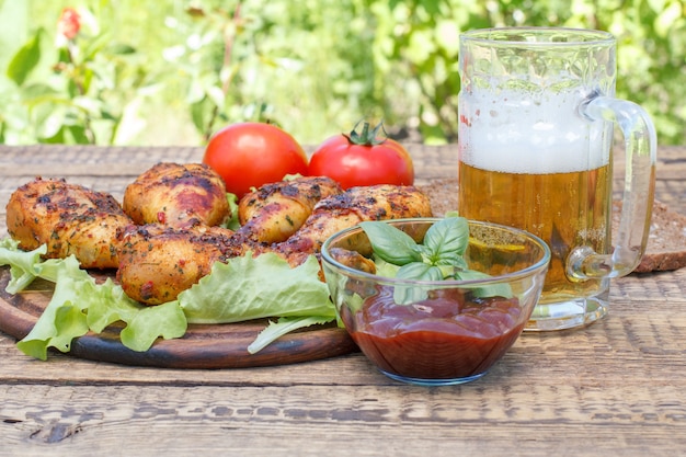 Cuisses de poulet grillées et feuilles de laitue sur planche à découper en bois, sauce tomate dans un bol en verre décoré de basilic vert, chope de bière en verre, tomates fraîches et pain noir