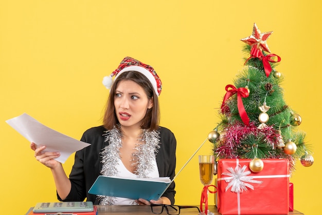 Curieuse charmante dame en costume avec chapeau de père Noël et décorations de nouvel an tenant un document au bureau sur isolé jaune