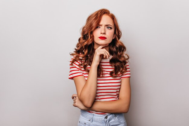 Curieuse dame européenne en tenue décontractée debout sur un mur blanc. Jolie femme aux cheveux longs posant.