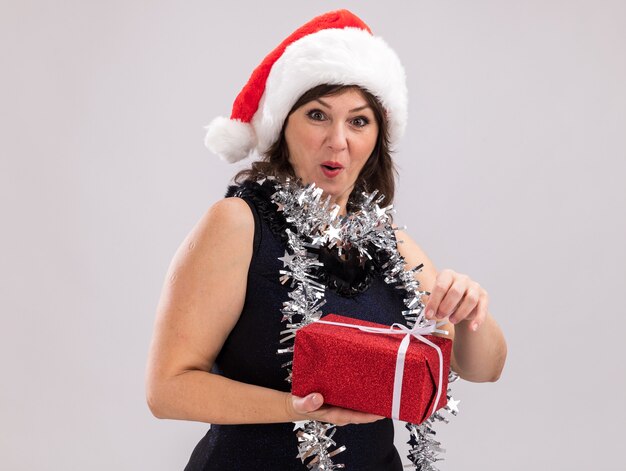 Curieuse femme d'âge moyen portant un bonnet de noel et une guirlande de guirlandes autour du cou tenant un paquet cadeau de noël saisissant un ruban regardant la caméra isolée sur fond blanc