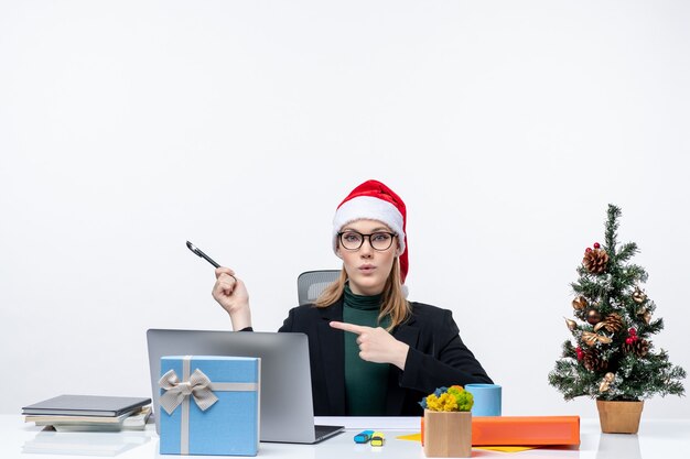 Curieuse femme blonde avec un chapeau de père Noël assis à une table avec un arbre de Noël et un cadeau dessus et pinte quelque chose sur fond blanc