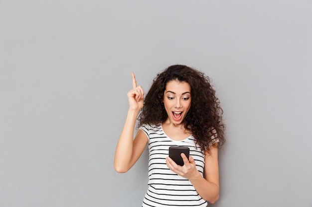 Curieuse femme regardant un téléphone portable en gardant l'index pointé vers le haut exprimant qu'elle a une idée ou une solution sur un mur gris