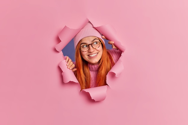 Photo gratuite curieuse femme rousse joyeuse concentrée de côté voit quelque chose de très agréable sourit largement coiffé d'un chapeau et de lunettes.