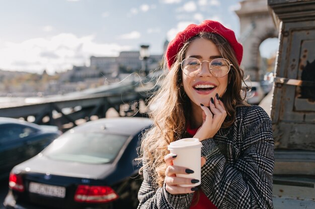 Curieuse fille blonde en élégant béret rouge posant avec le sourire sur fond flou en matin venteux