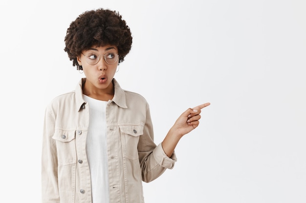 Curieuse et intriguée charmante fille garçon à la peau sombre à lunettes et chemise beige pointant et regardant à droite avec les lèvres pliées et l'expression intéressée étant impliquée dans la conversation