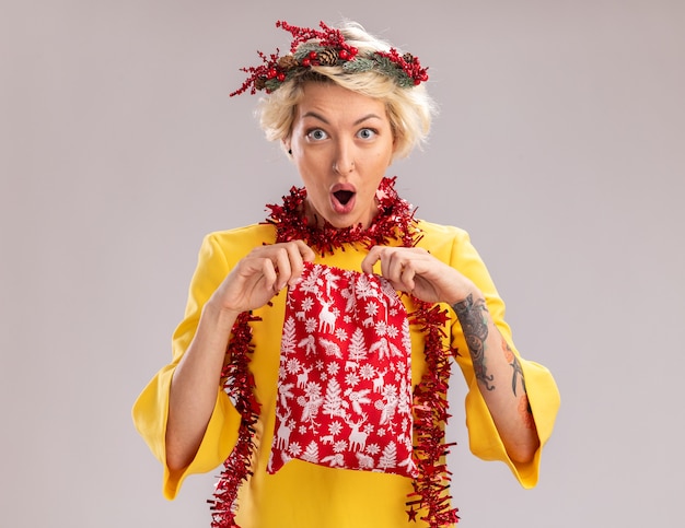 Photo gratuite curieuse jeune femme blonde portant une couronne de tête de noël et guirlande de guirlandes autour du cou tenant un sac de cadeau de noël en l'ouvrant en regardant la caméra isolée sur fond blanc