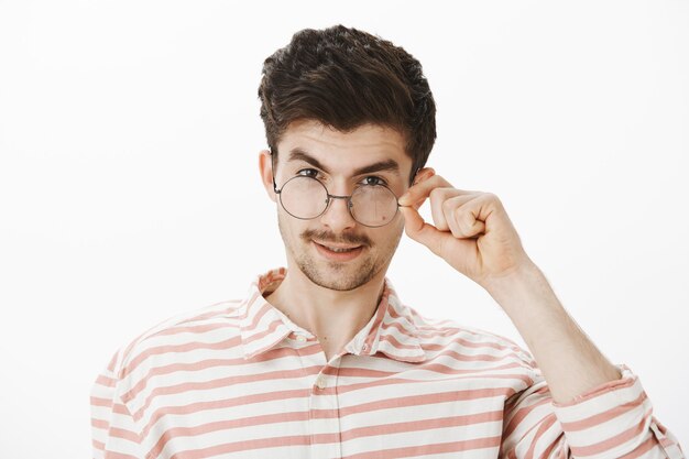 Curieux beau mec intéressant regardant une grande chose. Heureux homme séduisant avec barbe et moustache, à la recherche de dessous le front, décoller les lunettes, à la recherche avec intérêt sur mur gris