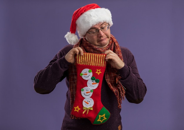 Photo gratuite curieux homme adulte portant des lunettes et un bonnet de noel avec une écharpe autour du cou tenant des bas de noël regardant à l'intérieur isolé sur un mur violet