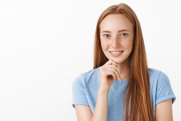 Curieux et intéressé modèle féminin rousse attrayant avec de jolies taches de rousseur souriant joyeusement tout en écoutant une histoire intéressante tenant la main sur le menton exprimant l'intrigue et la joie sur le mur gris