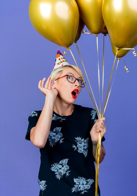 Curieux jeune femme de fête blonde portant des lunettes et une casquette d'anniversaire tenant des ballons à côté de faire ne peut pas vous entendre geste isolé sur mur violet