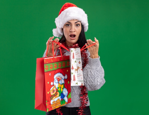 Photo gratuite curieux jeune fille caucasienne portant chapeau de noël et guirlande de guirlandes autour du cou tenant des sacs-cadeaux de noël ouvrant un isolé sur mur vert avec espace copie