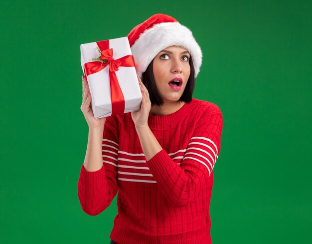 Curieux jeune fille portant un bonnet de Noel tenant un paquet cadeau près de la tête à l'écoute isolé sur un mur vert avec espace copie