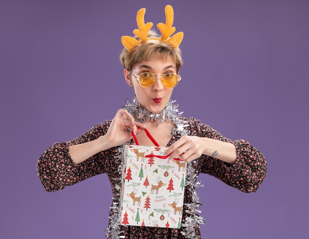 Curieux jeune jolie fille portant bandeau de bois de renne et guirlande de guirlandes autour du cou avec des lunettes tenant le sac de cadeau de Noël l'ouvrant en regardant la caméra isolée sur fond violet