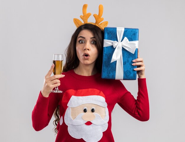 Curieux jeune jolie fille portant un bandeau de bois de renne et un pull du père Noël tenant un verre de champagne et un paquet cadeau près de la tête à la recherche