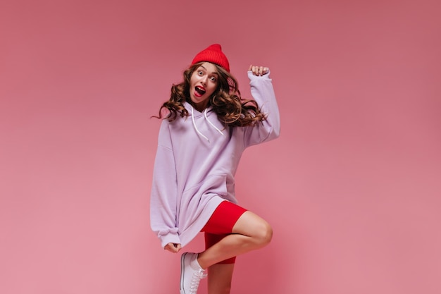 Photo gratuite curly brunette girl in red cycling shorts and purple hoodie sauts on isolated cool active woman in hat se déplace sur fond rose