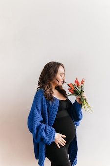 Curly femme enceinte heureuse en cardigan bleu détient des fleurs. charmante jeune fille brune en robe noire sent le bouquet de tulipes sur fond blanc.