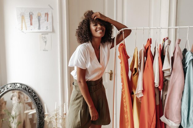 Curly jeune femme à la peau foncée en chemisier d'été blanc élégant et short kaki sourit sincèrement et s'appuie sur un cintre dans la salle blanche