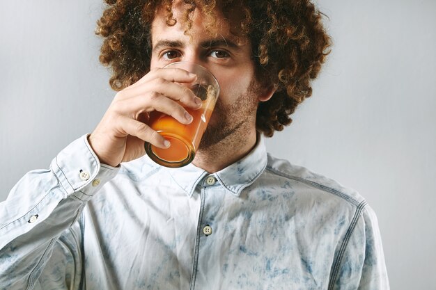 Curly jeune homme barbu en chemise de jeans blanc boit du jus naturel fraîchement pressé de carottes biologiques de la ferme.