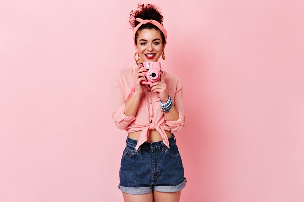 Curly lady en chemisier rose et short en jean sourit et tient une mini caméra sur un espace isolé.