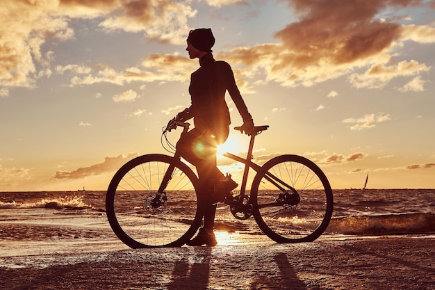 Photo gratuite cycliste féminine debout avec son vélo et profitant du coucher de soleil sur la côte de la mer.